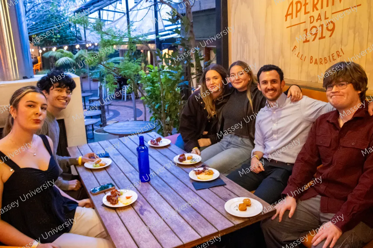 Six people share small plates around a wooden table at a plant-filled restaurant.