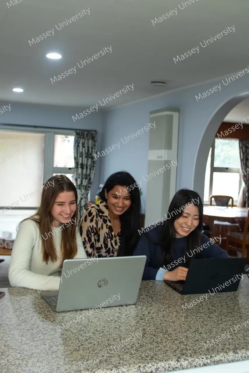 Three students smile as they work together on two laptops at a kitchen counter.