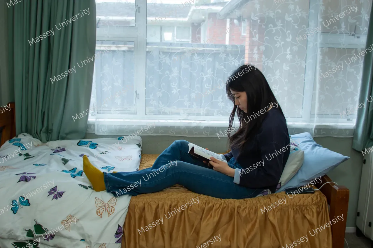 A student sits on a bed reading a book beside a large window.