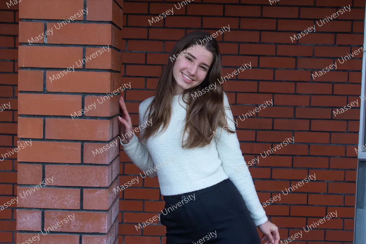 A smiling young woman in a white sweater leans against a red brick wall.