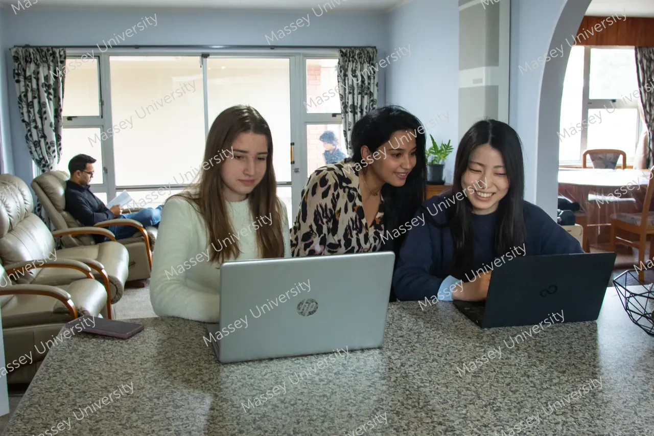 Three students work together on laptops at a table while another student sits on a sofa behind them.