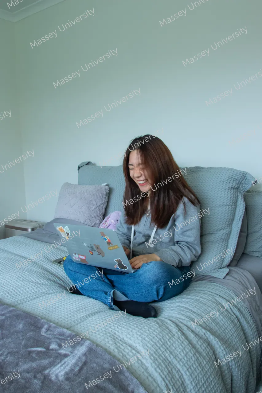 A smiling woman sits cross-legged on a bed, using a sticker-covered laptop.