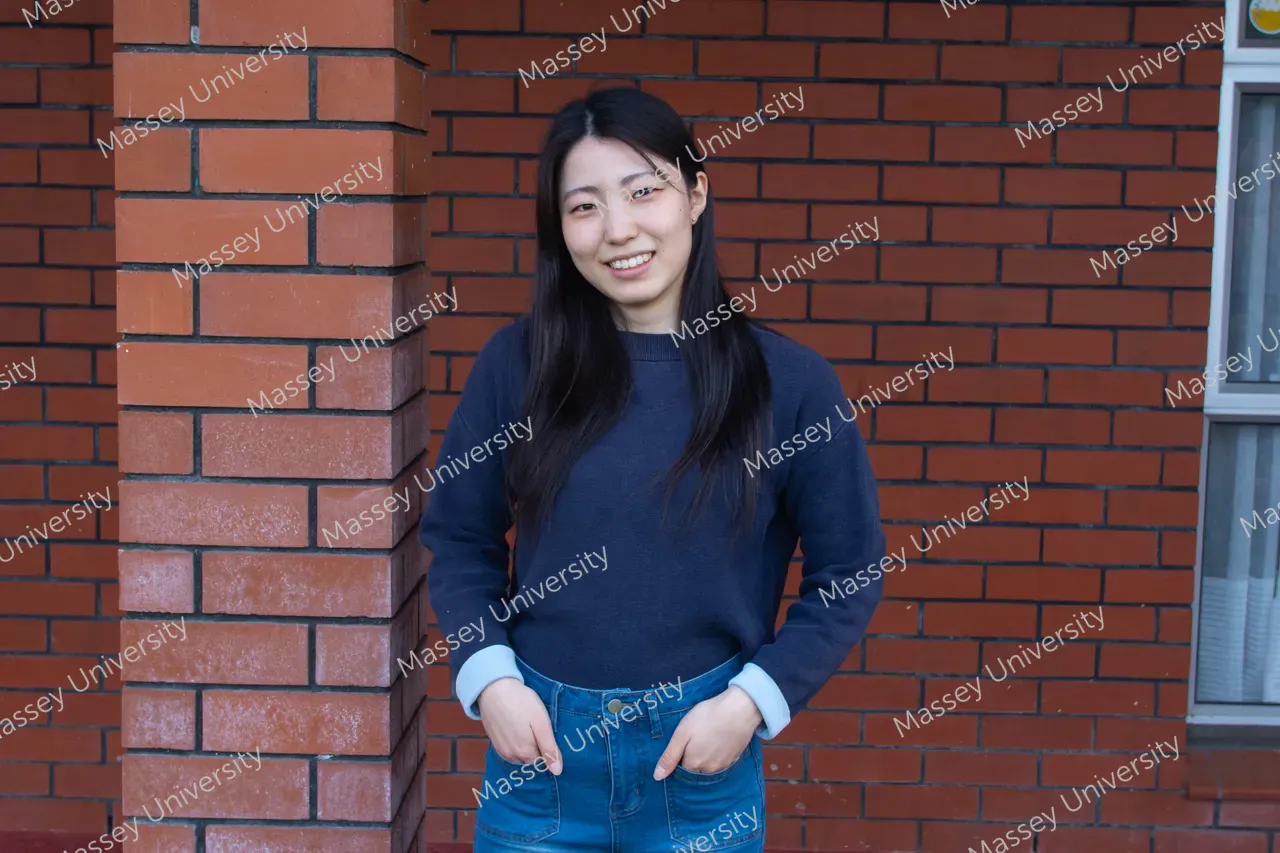 A smiling woman wearing a navy sweater and jeans stands against a brick wall.