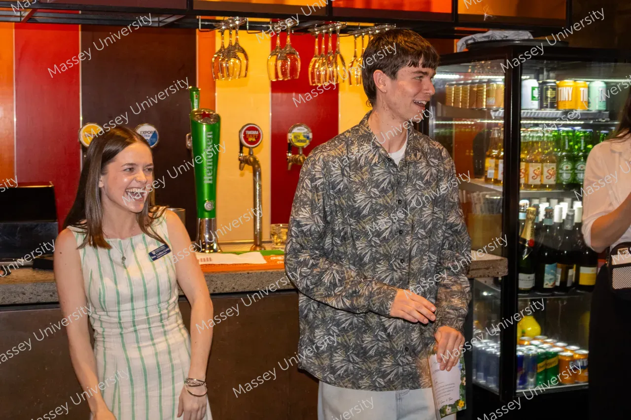 A woman and a man smile beside a bar counter with hanging glasses and a drinks refrigerator behind them.