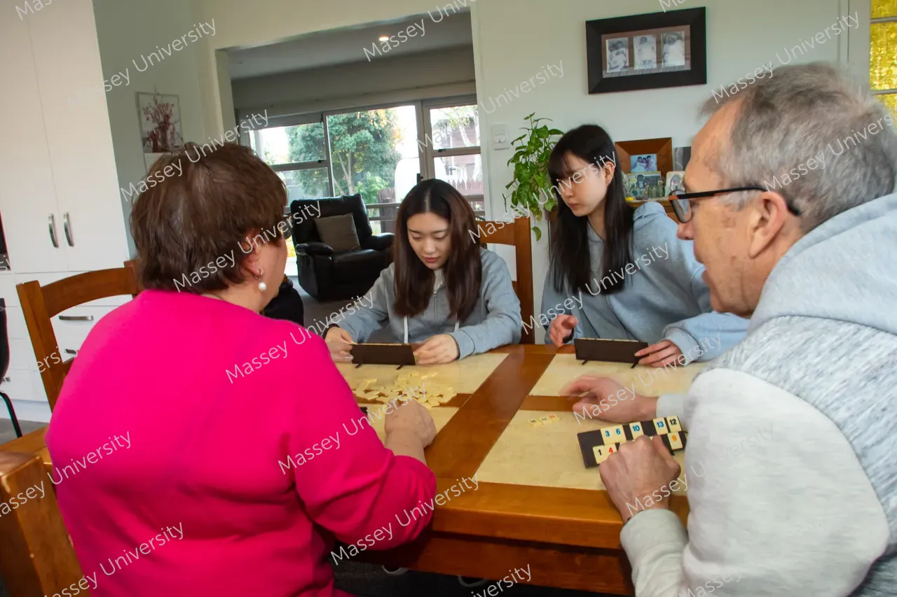 Two students and two older adults play a tile-based board game around a wooden table.